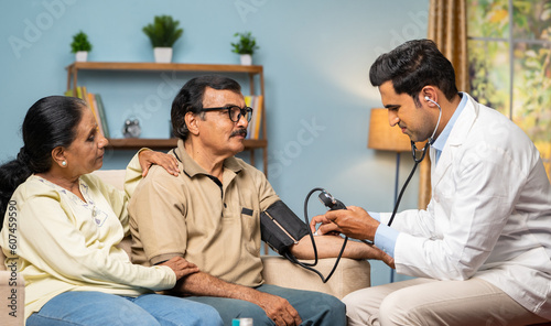 Indian doctor checking bp or blood pressure to senior man while sitting on sofa with wife at home - concept of home health checkup, medical treatement and relationship. photo