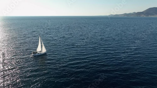 A white sailing yacht goes along the coast to the port on the horizon a smooth shot from a drone the rays of the sun are reflected on the sea surface