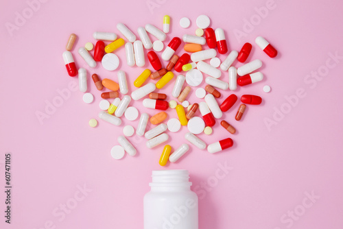 White bottle container with various colorful medication tablets and capsules on pink background. Concept of healthcare and medicine. Top view
