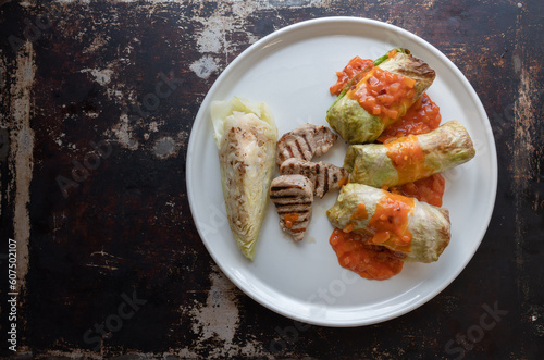 Cabbage rolls with grilled pork tenderloin, cooked cabbage and tomato sauce on a white plate and table, top view