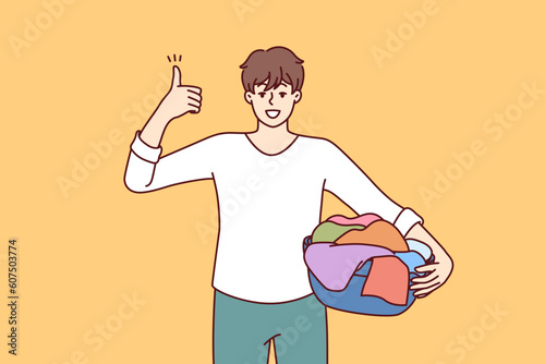 Man householder with laundry in basin shows thumbs up recommending quality laundry detergent. Guy getting ready to load textiles into washing machine setting positive example for peers