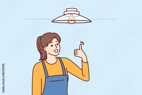 Woman electrician showing thumbs up after fixing chandelier or replacing light bulb under ceiling. Girl in overalls works as electrician efficiently performing tasks of setting up electricity