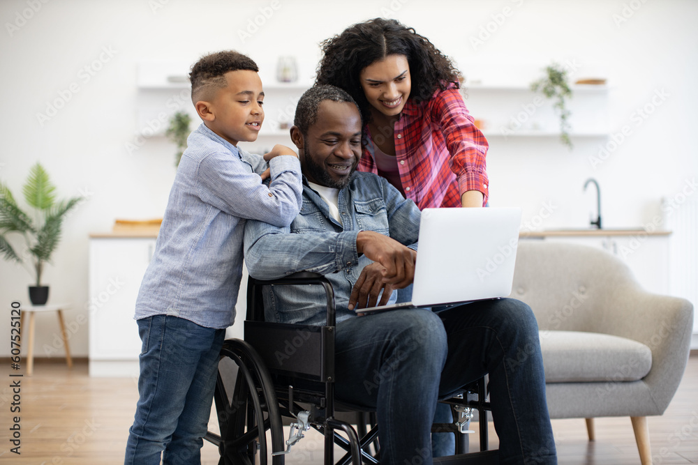 Cheerful multiracial man using wheelchair holding laptop on knees while pretty woman and funny boy standing behind. Loving family of three choosing movie for watching together on Sunday at home. Cheerful multiracial man using wheelchair holding laptop on knees while pretty woman and funny boy standing behind. Loving family of three choosing movie for watching together on Sunday at home.