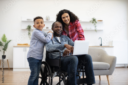 Cheerful multiracial man using wheelchair holding laptop on knees while pretty woman and funny boy standing behind. Loving family of three choosing movie for watching together on Sunday at home.
