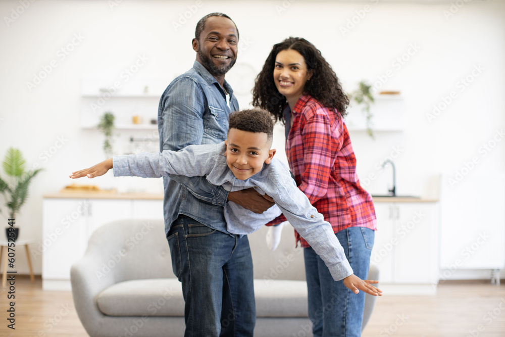 Cheerful multiracial child in casual clothes flying in parents' arms while pretending being pilot at home during weekend. Adventurous family of three changing rules of paper plane contest indoors. Cheerful multiracial child in casual clothes flying in parents' arms while pretending being pilot at home during weekend. Adventurous family of three changing rules of paper plane contest indoors.