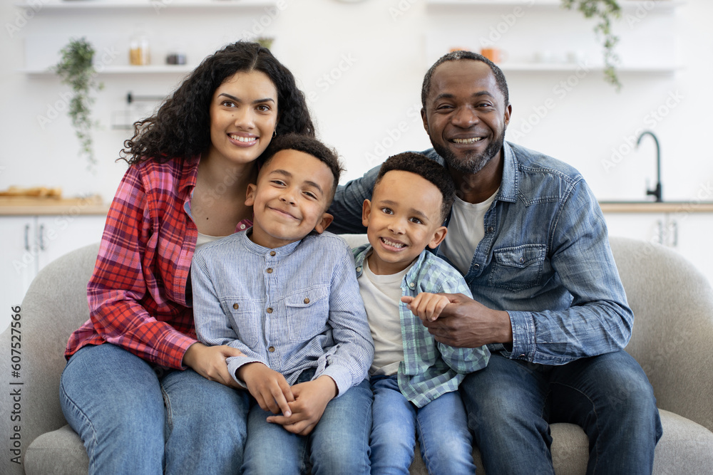 Beautiful multiethnic spouses cuddling smiling boys while making family portrait in lounge interior. Cheerful young adults showing care and affection to well-behaved sons at leisure indoors. Beautiful multiethnic spouses cuddling smiling boys while making family portrait in lounge interior. Cheerful young adults showing care and affection to well-behaved sons at leisure indoors.