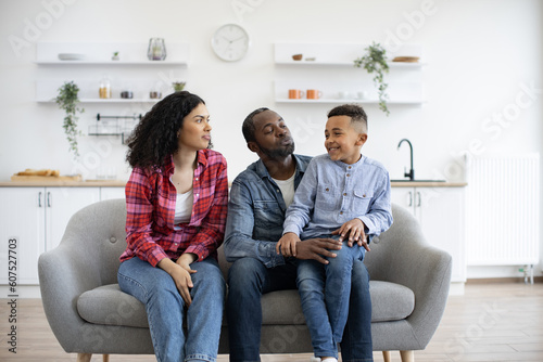 Inquisitive african american father holding son on knee while attentive mother asking about birthday presents. Inventive married couple planning family celebration while talking in living room.