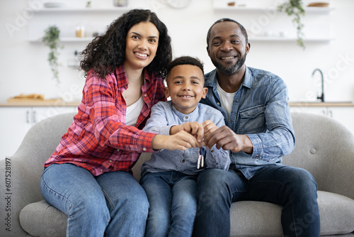 Happy multiracial adults and little kid in denim outfit holding keys in hands together while sitting on cozy couch indoors. Delighted owners of new apartment celebrating purchase on moving day.