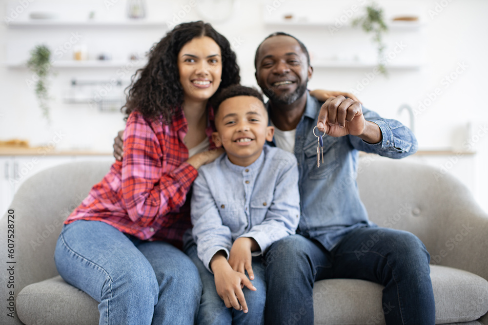 Proud african american father showing keys from new property while cuddling happy wife and playful son. Cheerful family of three in jeans relaxing on cozy sofa of newly bought modern apartment.