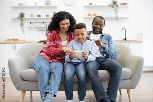 Inquisitive multiracial couple pointing fingers on phone screen held by calm little boy while relaxing on couch in lounge. Helpful parents giving advice about online video game after tasty dinner. photo