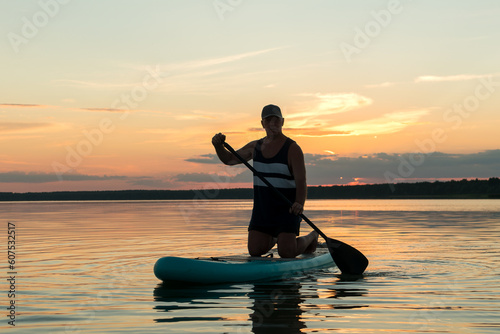 A man on his knees on a SUP board with an oar swims in the calm water of the lake against the backdrop of a pink sky reflection in the water.