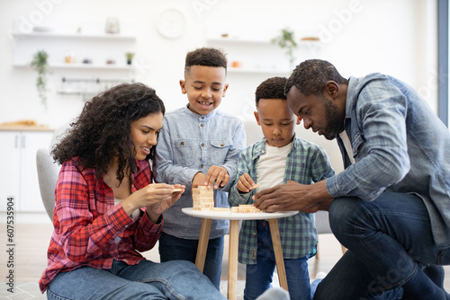 Close up view of four-person family playing board game with wooden blocks on small table in living room. Happy people being engaged in enjoyable activity showing importance of love and care.