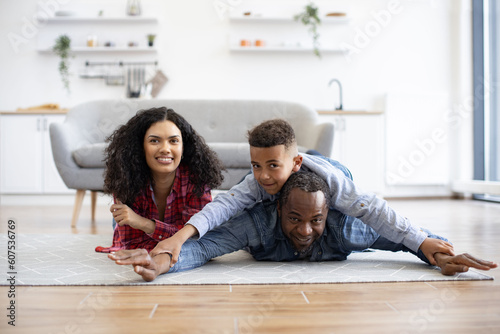 Healthy african american father holding son on back while staying with beautiful wife on kitchen floor. Affectionate dad showing swimming style to inquisitive boy while mom smiling happily aside.