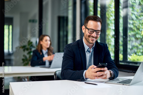 A smiling businessman with glasses on typing on a mobile while working at the office.