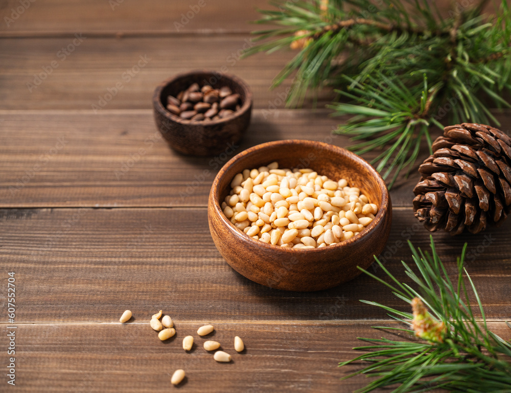 Pine nuts in a bowl and a handful of unpeeled nuts on a brown wooden background with a branch of pine needles. Healthy diet snack. Pine nuts in a bowl and a handful of unpeeled nuts on a brown wooden background with a branch of pine needles. Healthy diet snack.