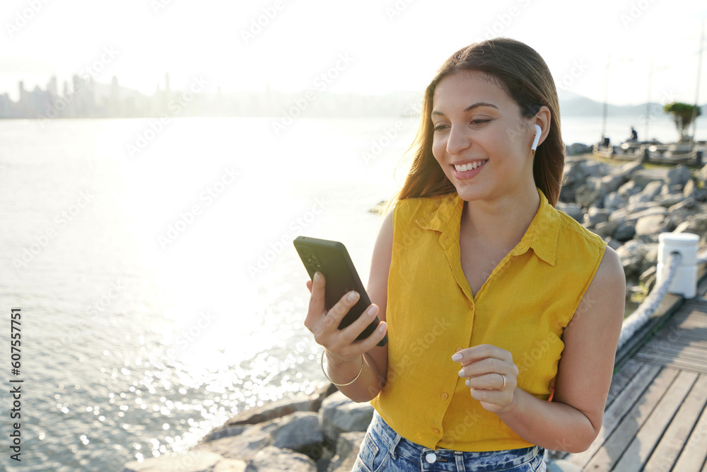 Portrait of cheerful attractive young woman wearing earphones using mobile phone outdoor with sunset in background. People and technology outdoor leisure activity. Portrait of cheerful attractive young woman wearing earphones using mobile phone outdoor with sunset in background. People and technology outdoor leisure activity.