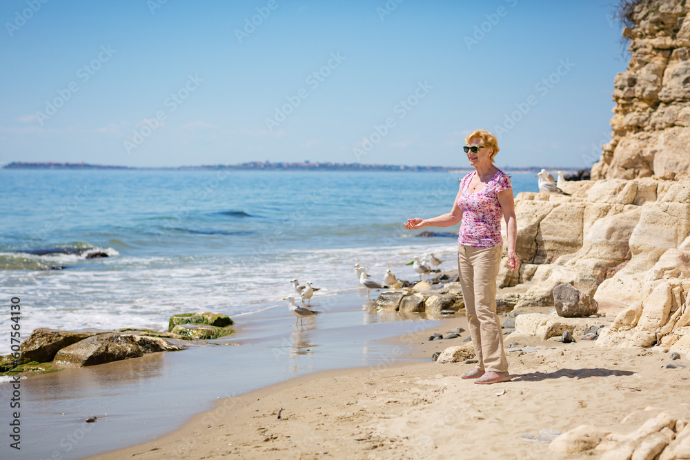 Elderly woman in sunglasses feeds seagulls on the beach and laughs. Happy retirement life