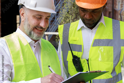 Multiracial team of competent builders in protective helmets working together on project plan outdoors. Adult caucasian foreman with clipboard recording step-by-step actions.