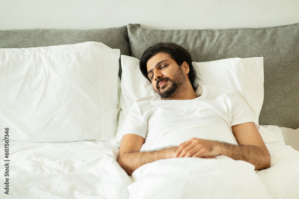 Millennial middle eastern guy sleeping, lies on white bed with empty space in bedroom interior Millennial middle eastern guy sleeping, lies on white bed with empty space in bedroom interior