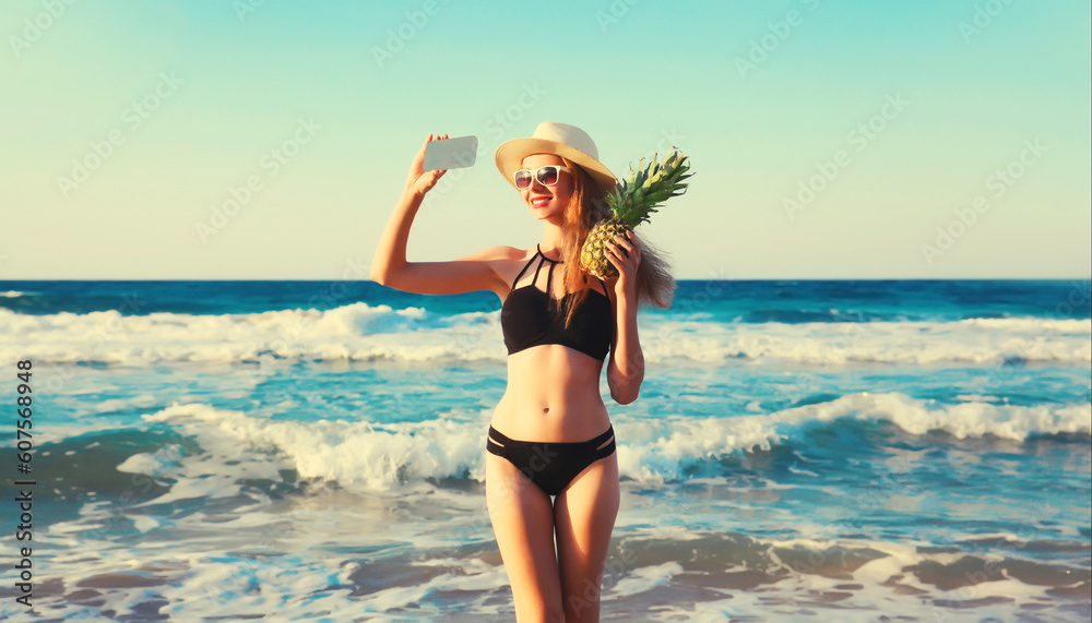 Summer vacation, portrait of happy smiling young woman holding pineapple taking selfie with smartphone wearing bikini swimsuit and straw hat on the beach on sea coast with waves background