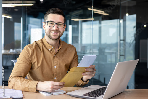 Portrait of a young business man working in the office at a desk with a laptop and holding an envelope with a letter and documents in his hands. He looks at the camera with a smile