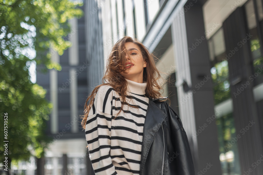 Beautiful young girl model in a fashionable outfit with a striped sweater and a leather jacket walks in a modern city Beautiful young girl model in a fashionable outfit with a striped sweater and a leather jacket walks in a modern city