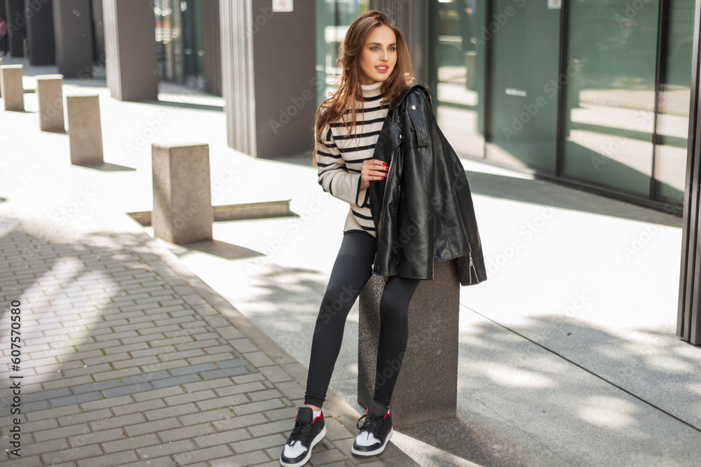 Fototapeta premium Fashionable beautiful happy girl in stylish clothes with leather jacket, sweater, leggings and sneakers sits in a sunny city near a modern building