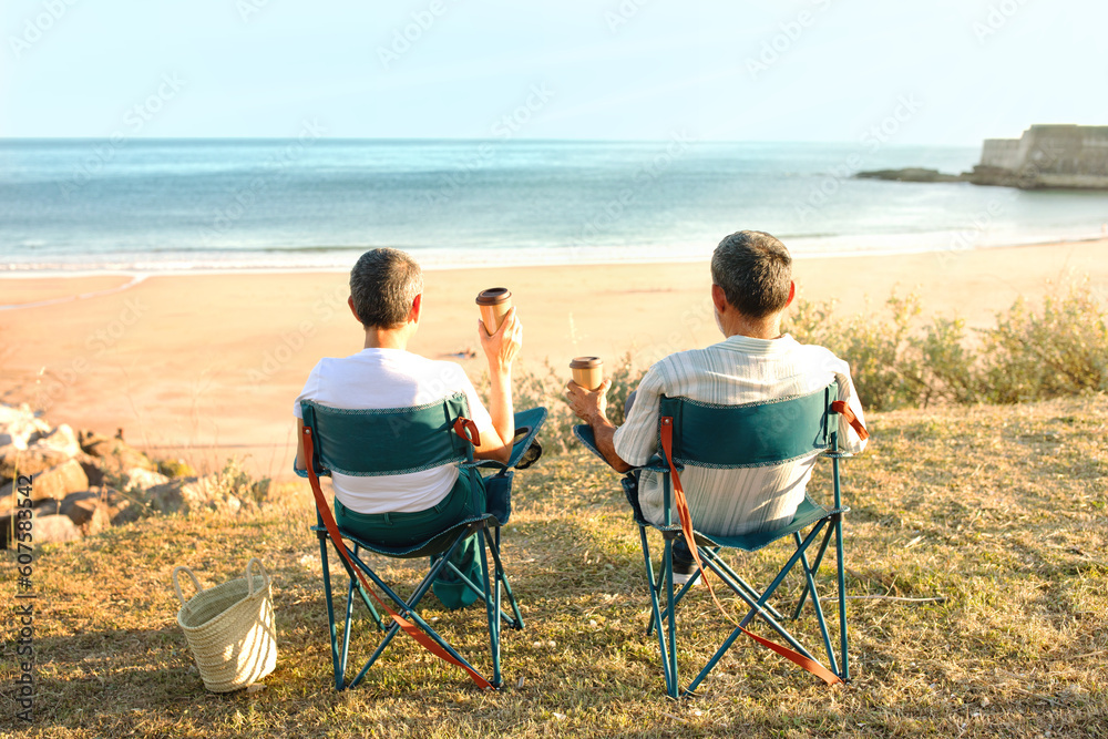 Senior Couple Sitting In Chairs Drinking Coffee Near Sea Outside