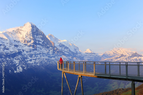 A visitor in red jacket standing on the view platform Cliff Walk at the First, Gwindelwald in the morning sunrise hours against the snow mountains