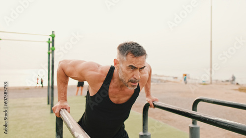 Middle-aged muscular man in sportswear pulls himself up on uneven bars on sea background