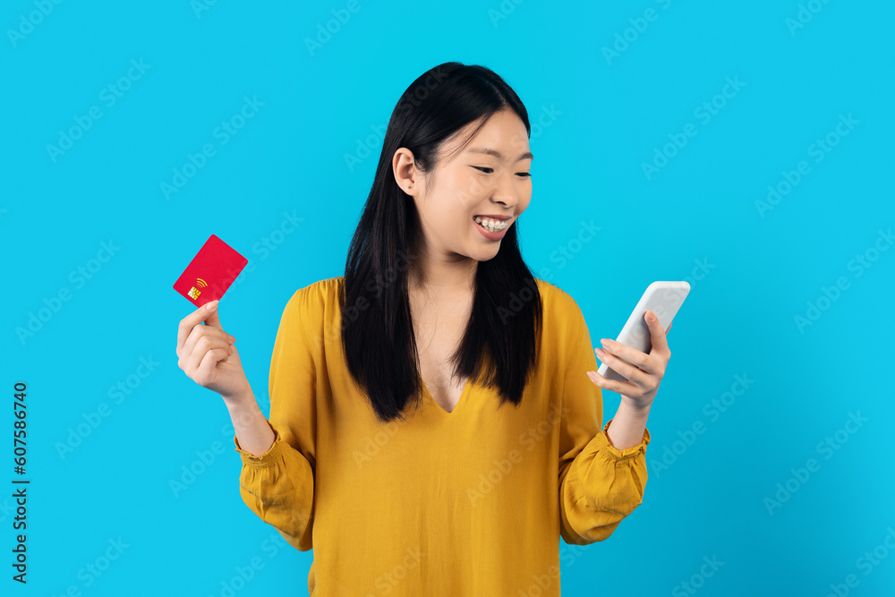 Cheerful asian woman holding smartphone and bank card on blue Cheerful asian woman holding smartphone and bank card on blue