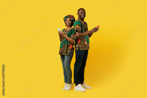 Happy black couple in african costumes using smartphones on yellow