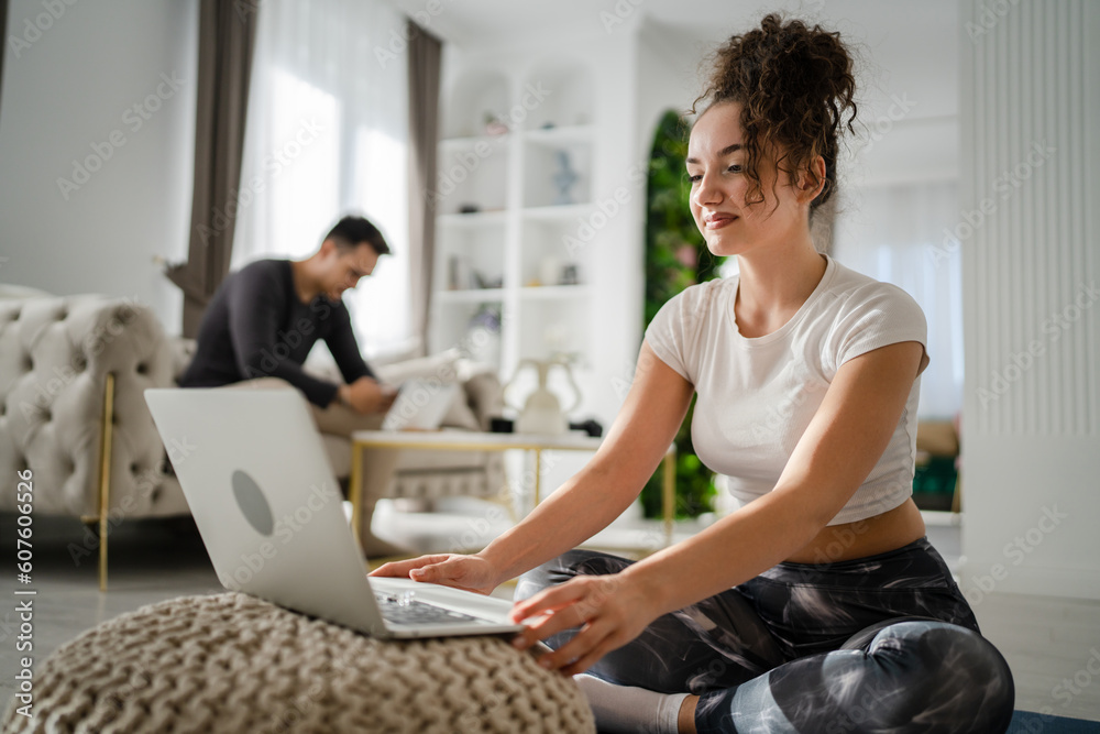 Fototapeta premium Woman young adult caucasian female sitting on the floor at home working on laptop computer browse internet online while her husband or boyfriend is sitting in the background leisure family concept