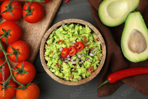 Bowl of delicious guacamole and ingredients on grey wooden table, flat lay
