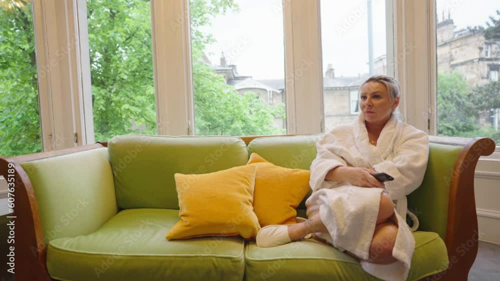 Woman sits on green sofa wearing dressing gown and using remote control Woman sits on green sofa wearing dressing gown and using remote control
