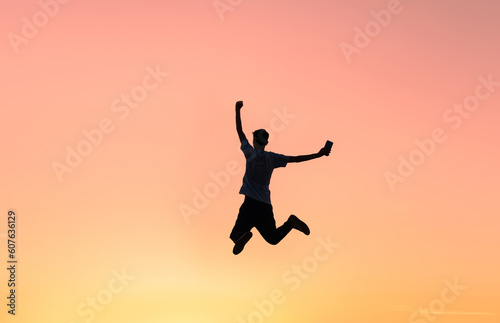 Silhouette of a man holding a smartphone jumping up to celebrate success  with sunset sky in the background. Motivation to achieve success.