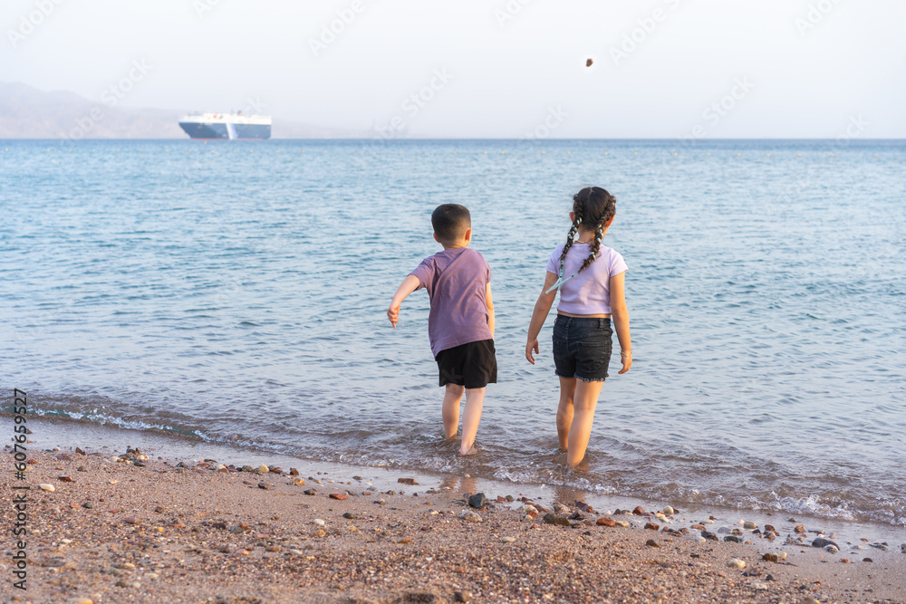Two young children playing on a pebble beach and throwing stones in the water.