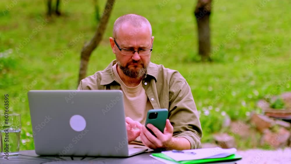Serious bearded man wearing glasses using laptop and mobile phone while sitting in the garden. Male freelancer working remotely outdoors. Digital nomad concept. Serious bearded man wearing glasses using laptop and mobile phone while sitting in the garden. Male freelancer working remotely outdoors. Digital nomad concept.