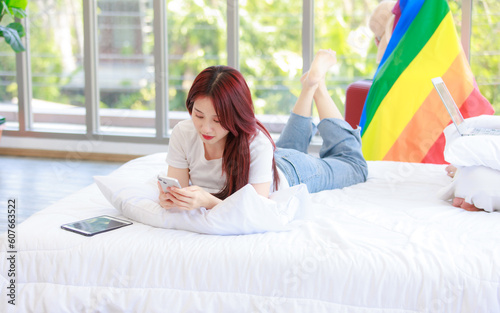 Asian young happy cheerful female LGBTQ lesbian lover sitting on bed in bedroom smiling using smartphone and tablet computer browsing surfing internet online with rainbow flag photo