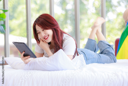 Asian young happy cheerful female LGBTQ lesbian lover laying lying down crossed legs on bed in bedroom smiling using smartphone and tablet computer browsing surfing internet online with rainbow flag photo