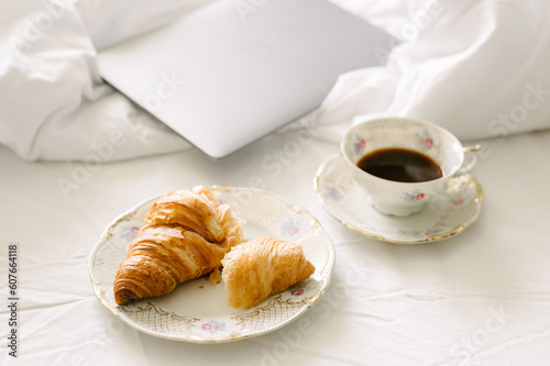 Overhead view of drink served in cup by smart computer laptop  croissant on bed.