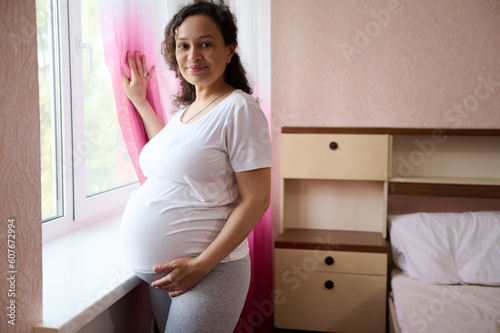 Portrait of young happy pregnant woman wearing white t-shirt, standing at window in the home bedroom, caressing touching her belly in late pregnancy, smiling looking at camera. Motherhood. Maternity © Taras Grebinets
