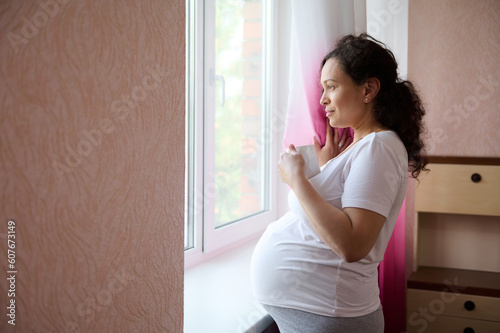 Beautiful pregnant woman future mother holds a cup of healthy tea, looking dreamily out the window at home, enjoying her pregnancy and maternity lifestyle. Motherhood. Expectation 40 week Trimester 3d © Taras Grebinets