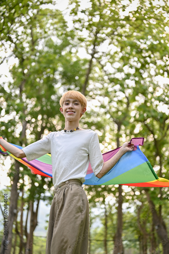 A happy  young Asian gay man with an LGBT rainbow flag standing in the park. Pride parade