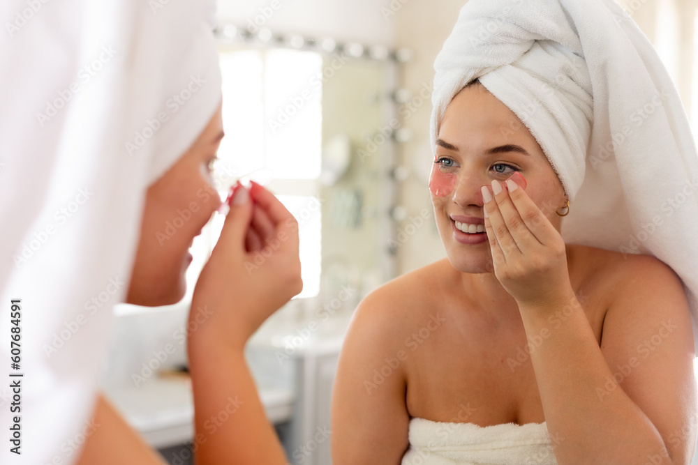 Caucasian plus size woman with towel wrapped on head applying eye patch while looking in mirror Caucasian plus size woman with towel wrapped on head applying eye patch while looking in mirror