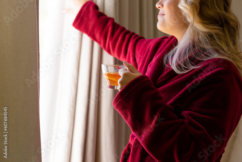 Midsection of plus size caucasian woman dressed in bathrobe holding tea cup opening window curtain photo