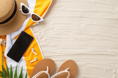Get your tan on. Accessories laid out from top view: glasses, cap, smartphone, earbuds, flip-flops, towel, shell, palm leaves. Sandy beach scene with an empty space for text or promotion