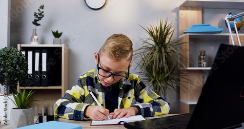 Front view of good-looking careful 10-aged modern boy in glasses which writing down the notes into notebook from computer photo