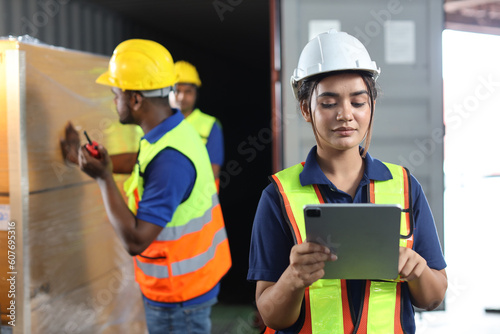 Group of warehouse workers with hardhats and reflective jackets using tablet, walkie talkie radio and cardboard while controlling stock and inventory in retail warehouse logistics, distribution center