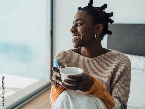 Smiling woman with Afro hairstyle sitting on bed holding coffee cup photo